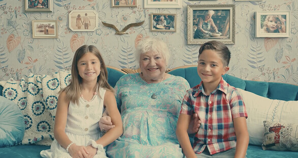 Smiling elderly woman sitting on a couch with her two grandchildren, surrounded by family photos in a warmly decorated living room.